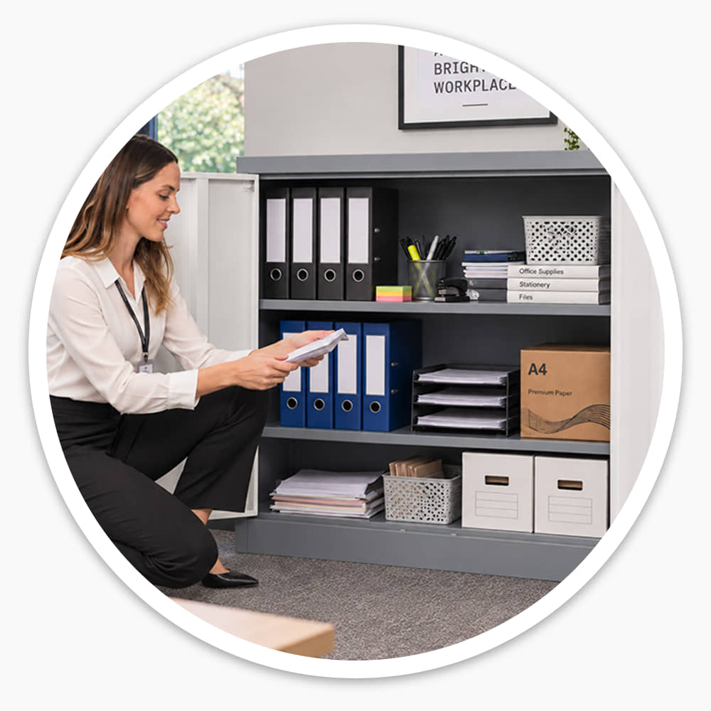 Woman filing documents in an open mid grey lockable cabinet holding box files in an office