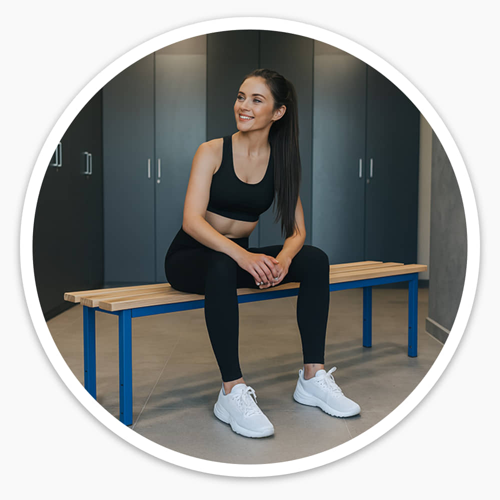 Woman in gym kit sitting on the 1500mm ash slatted cloakroom bench in a changing room lined with grey lockers