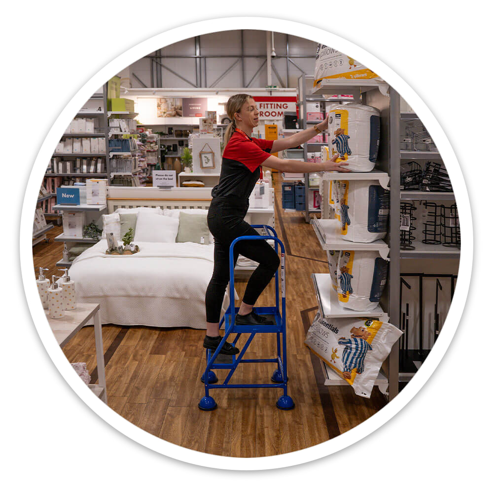 Retail assistant carrying stock on the 2 tread mobile safety steps in a shop display aisle