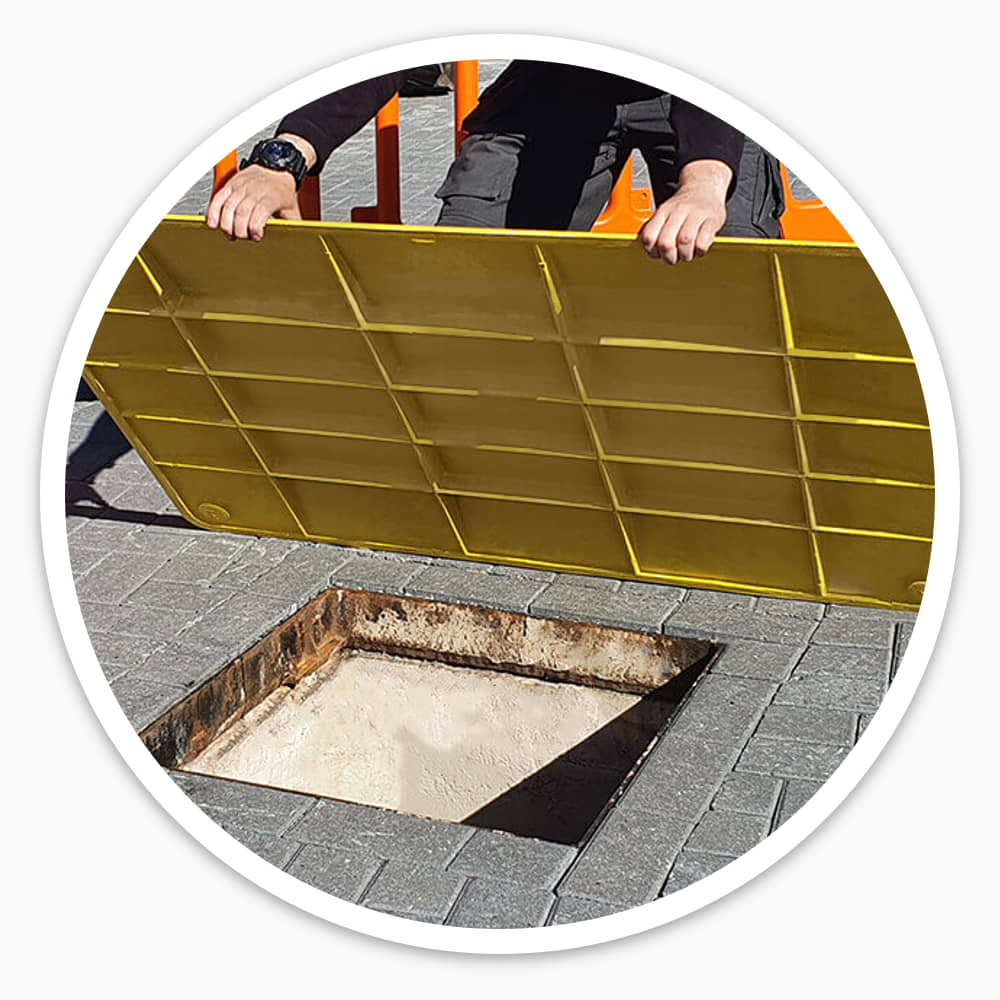 Worker lifting a Pedestrian Hole Cover to show its ribbed underside above an open hole in block paving