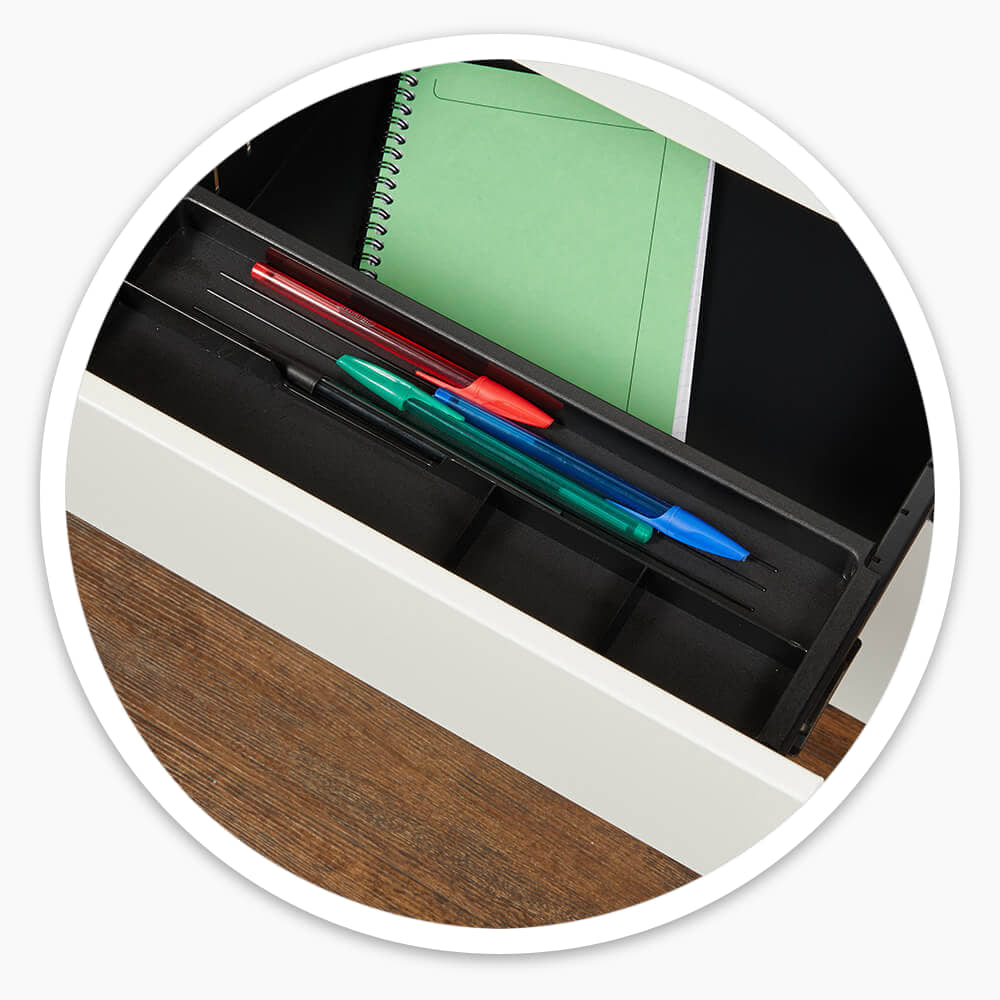Open top drawer of the white 3 drawer mobile steel pedestal, with pens in a black tray and a green notebook