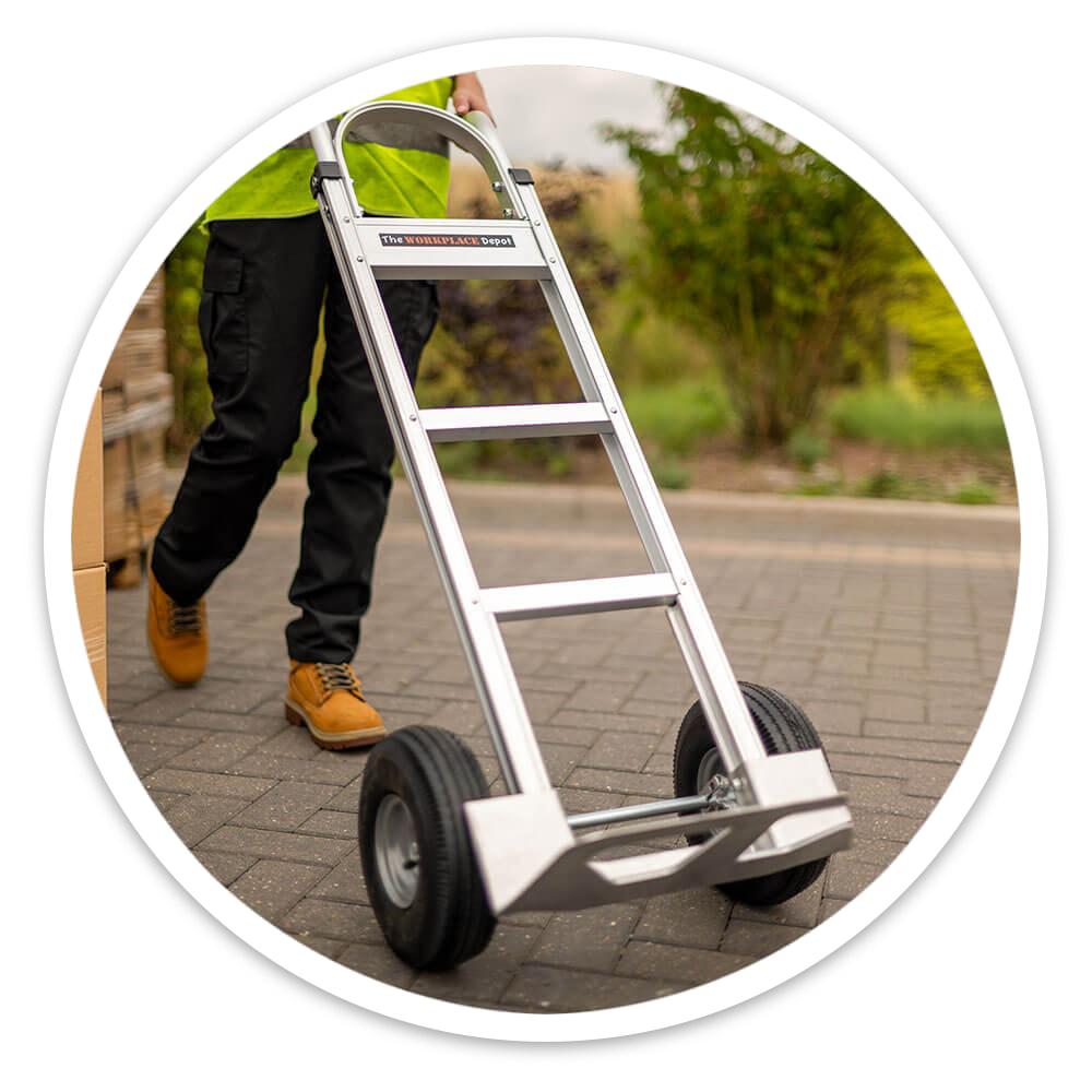 Worker in hi-vis wheeling an empty aluminium sack truck with pneumatic tyres across a block-paved yard beside boxes