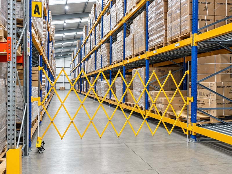Yellow steel expanding aisle barrier on its braked wheels, partly open across a warehouse aisle between pallet racking